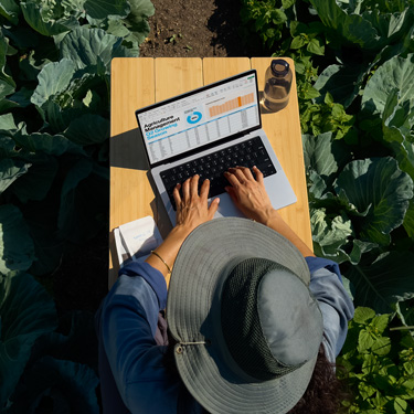 Bovenaanzicht van een vrouw met een hoed met brede rand die aan een tuintafel werkt aan een zakelijke presentatie op een MacBook.