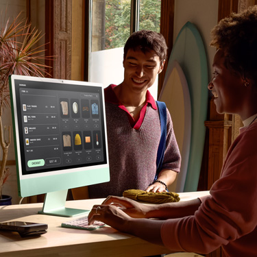 Een medewerker zit achter de toonbank in een winkel die surfplanken en andere artikelen verkoopt en gebruikt een iMac om een aankoop voor een klant af te ronden.
