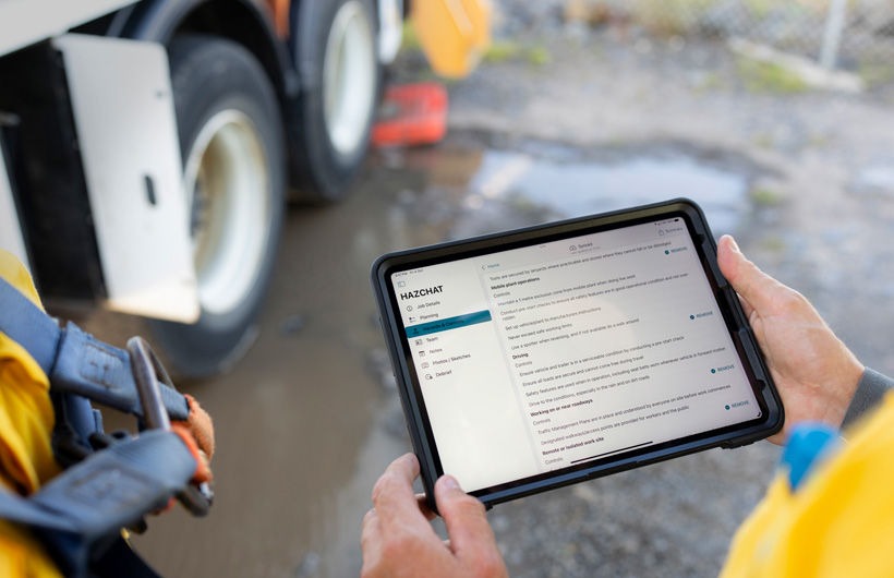Hender holder en iPad med HazChat-appen åpen, og på skjermen vises Job Details, Planning, Hazards og Controls. I bakgrunnen står en person ved en søledam på arbeidsstedet.