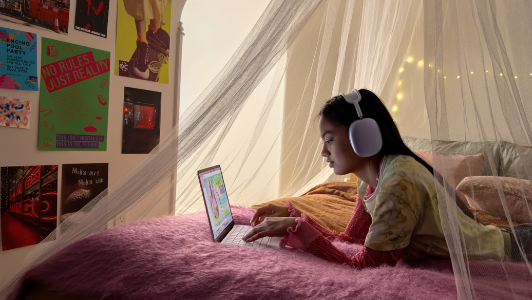A young person in their bedroom wearing headphones uses MacBook Neo, blush color, to book a nail appointment