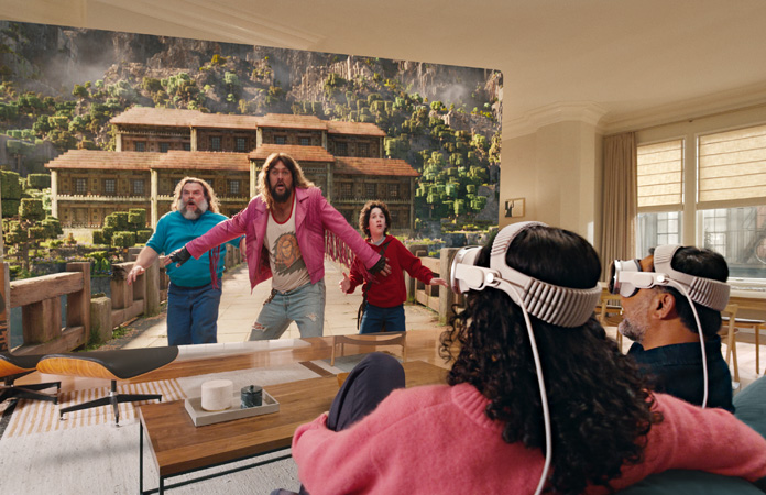 Deux personnes assises côte à côte sur un divan, portant un Apple Vision Pro et regardant Minecraft, le film, sur un écran qui s’étend d’un bout à l’autre de la pièce